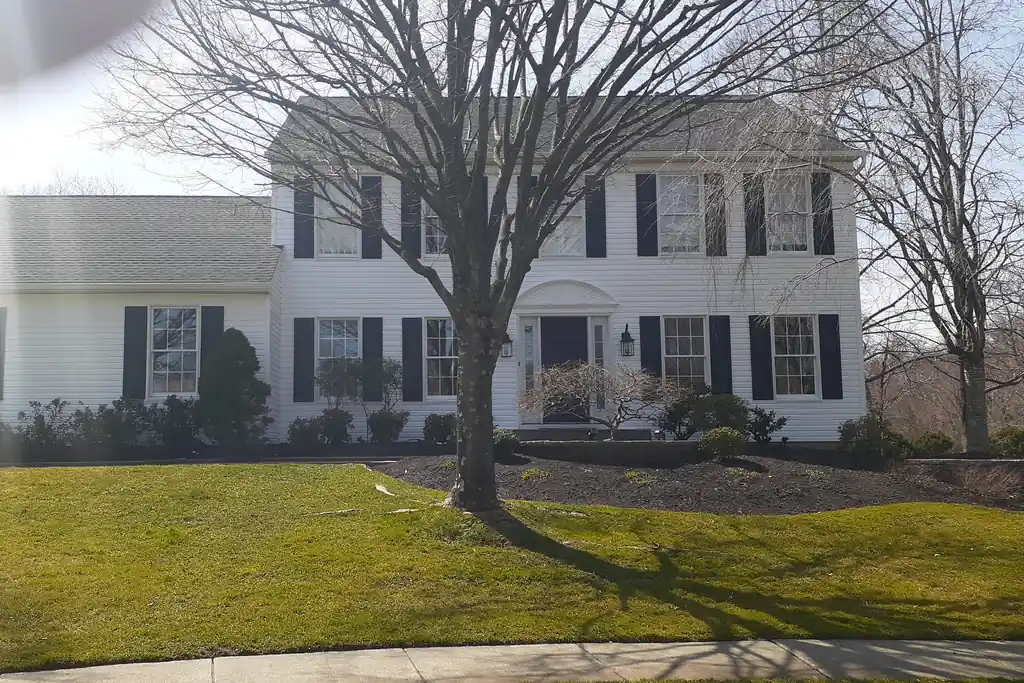 A white two-storey colonial house with black shutters and a grey shingle roof, seen from the street across a front lawn with a bare tree.