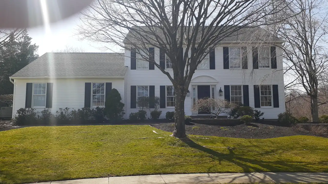 A white two-storey colonial house with black shutters and a grey shingle roof, seen from the street across a front lawn with a bare tree.