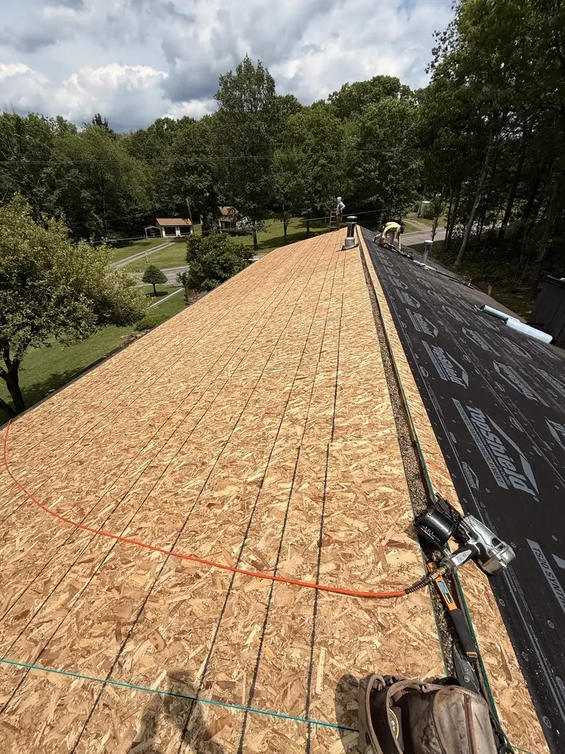 Roof stripped to bare decking with chalk lines snapped across it, underlayment rolled out on the far slope, a nail gun and air hose in the foreground and workers at the ridge.