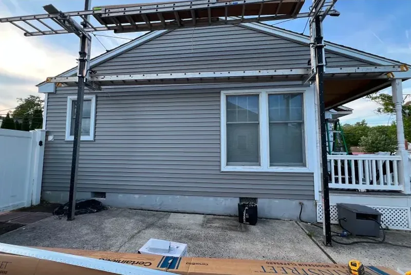 Gray vinyl siding part-installed on a single-story home, with a pergola frame overhead and a carton of MainStreet clapboard siding on the ground.