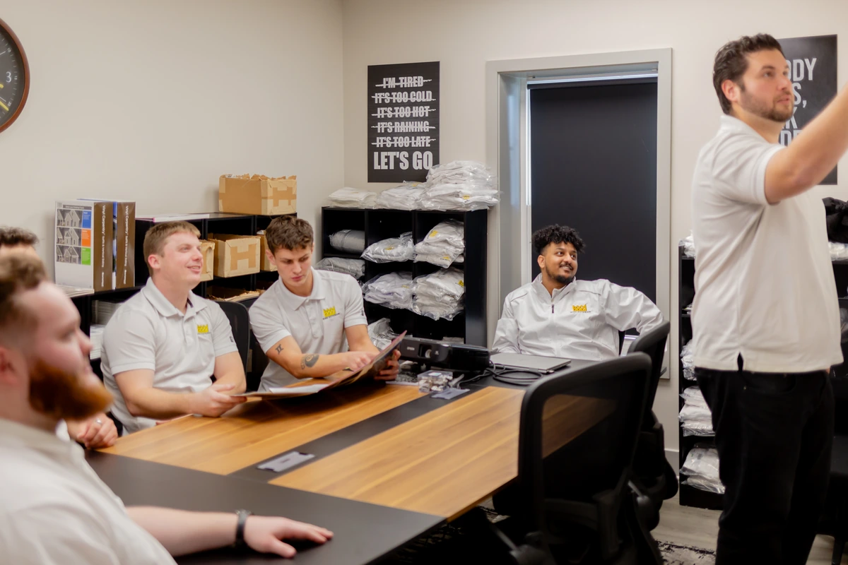 Six people in branded white shirts around a conference table in a meeting room, one standing and speaking to the group.
