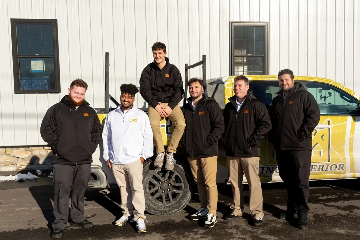 Six people in branded work jackets standing in front of a wrapped contractor pickup truck outside a metal-clad building in winter.
